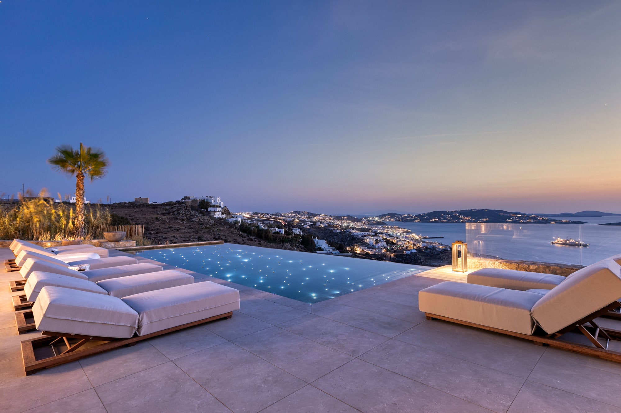 Outdoor pool area with lounge chairs overlooking a coastal town at dusk.