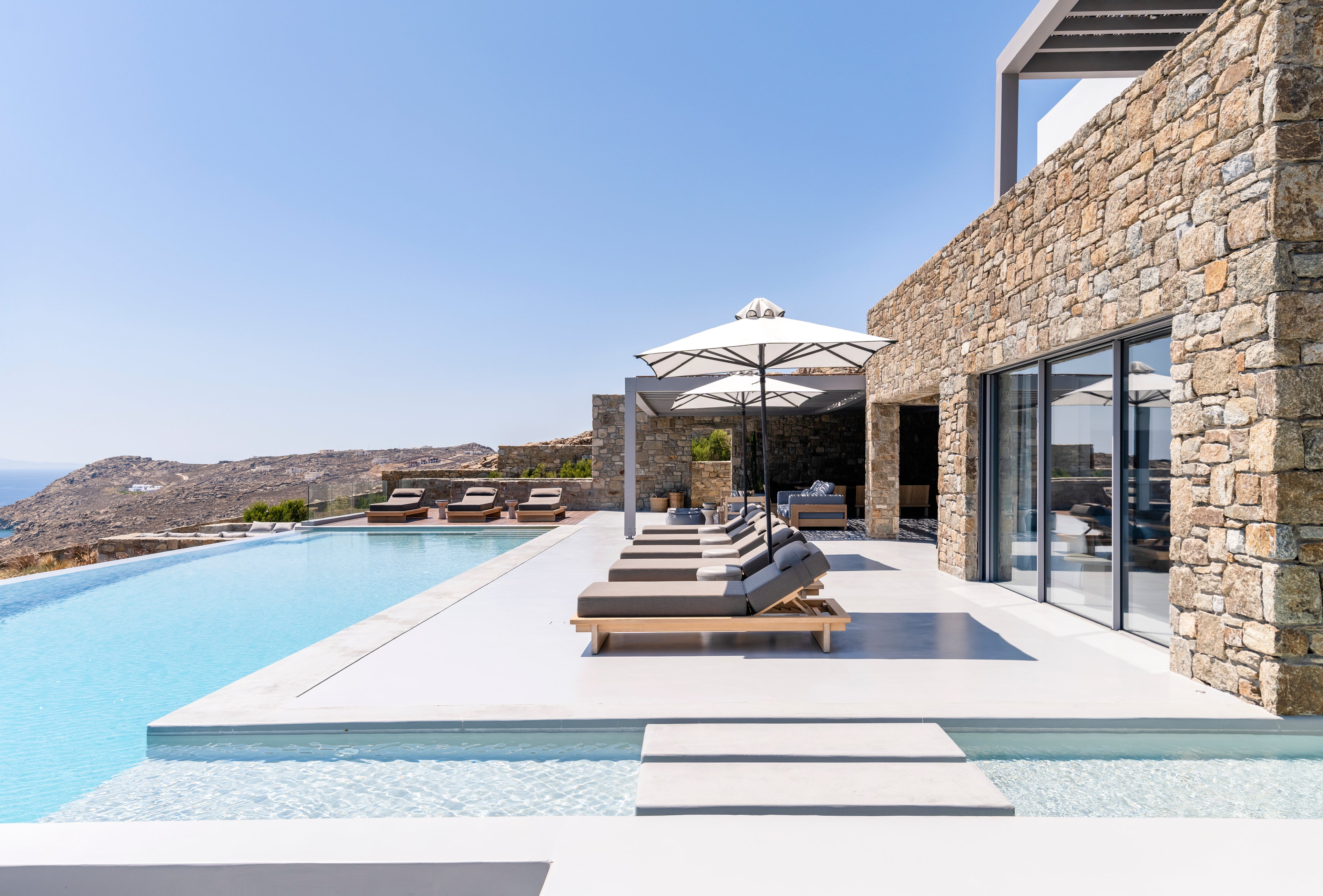 Modern outdoor pool area with lounge chairs, umbrellas, and stone wall.