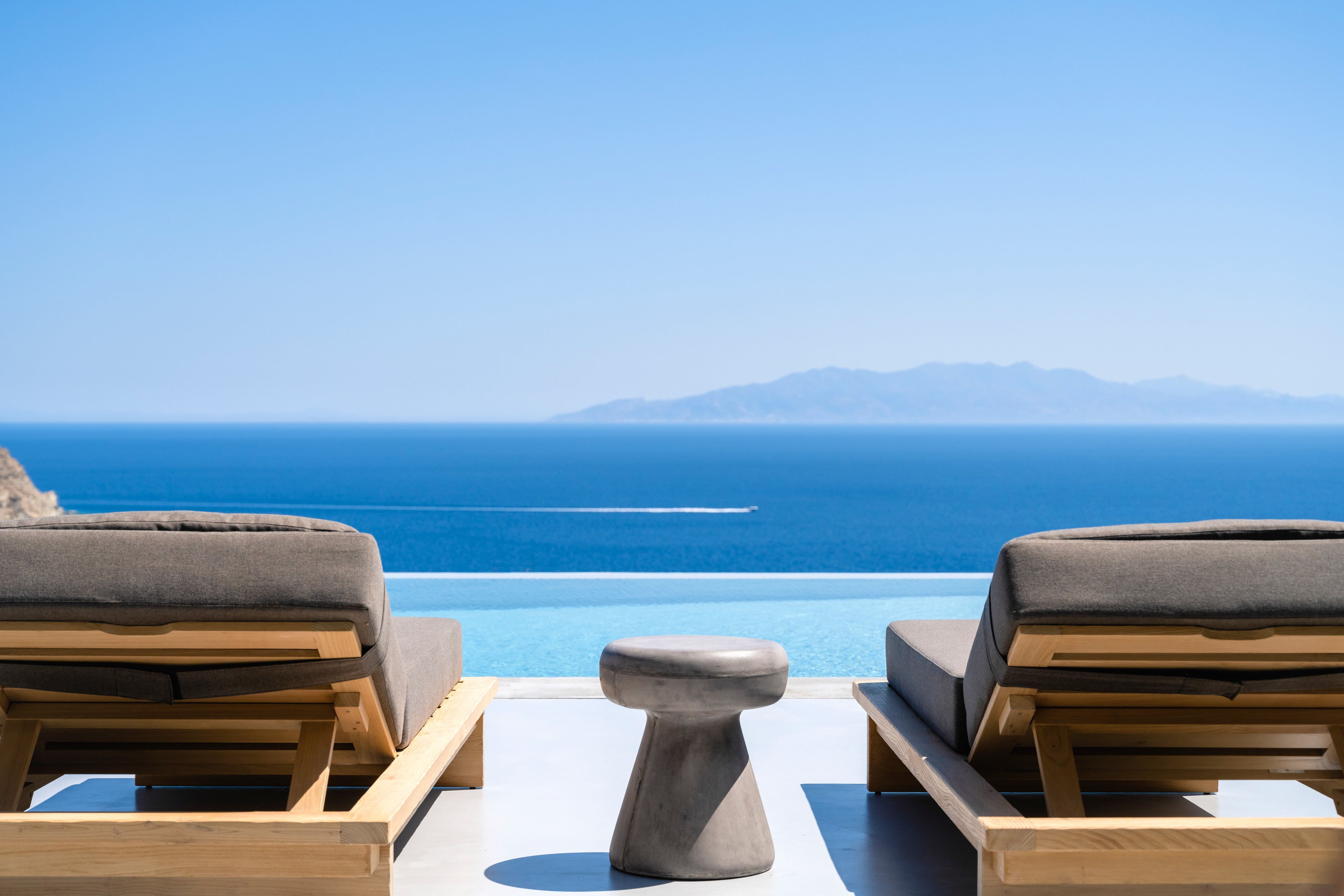 Two wooden lounge chairs with cushions on a balcony overlooking the ocean and mountains.