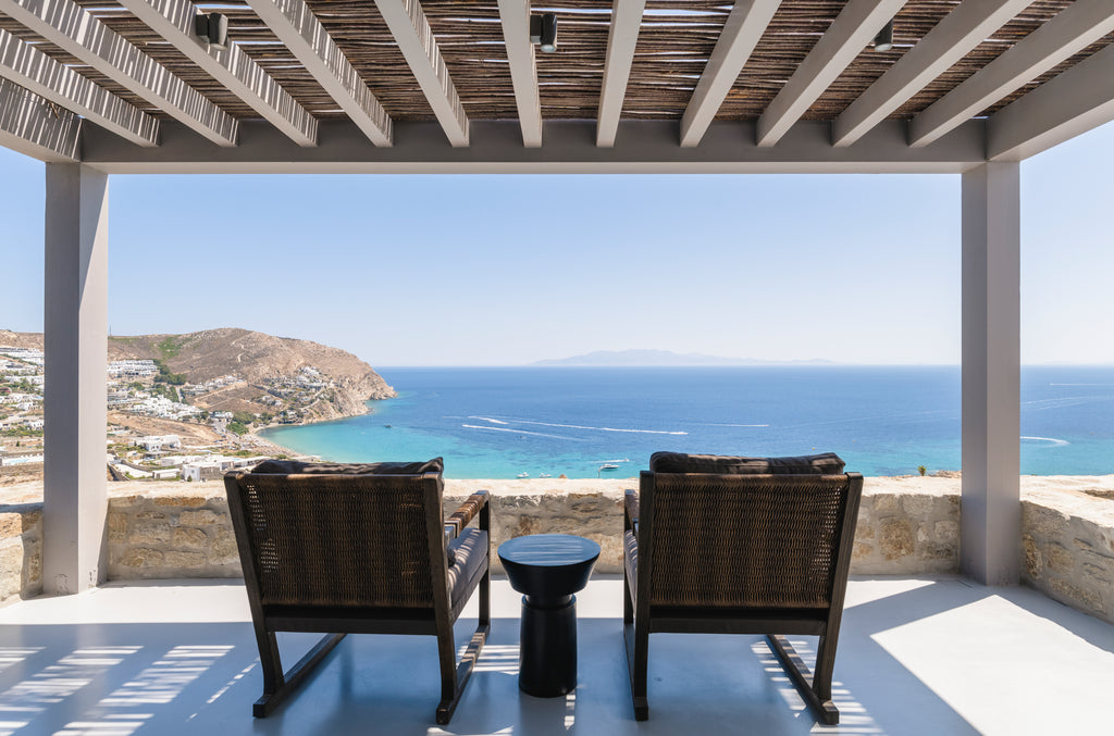 Two wicker chairs and a small table on a balcony with a scenic view of the ocean and coastline.