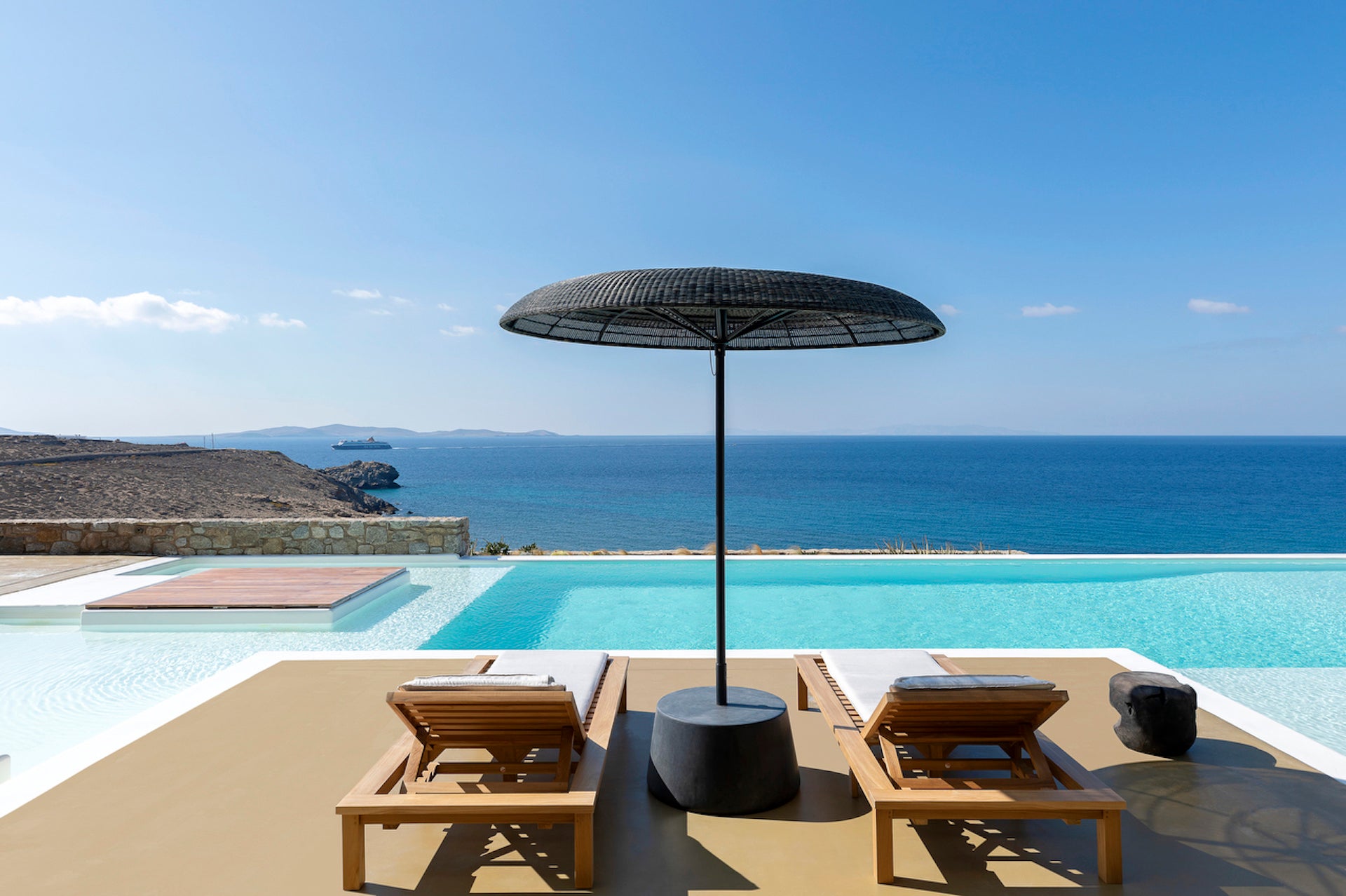 Two wooden lounge chairs under a large black umbrella by an infinity pool with ocean view.