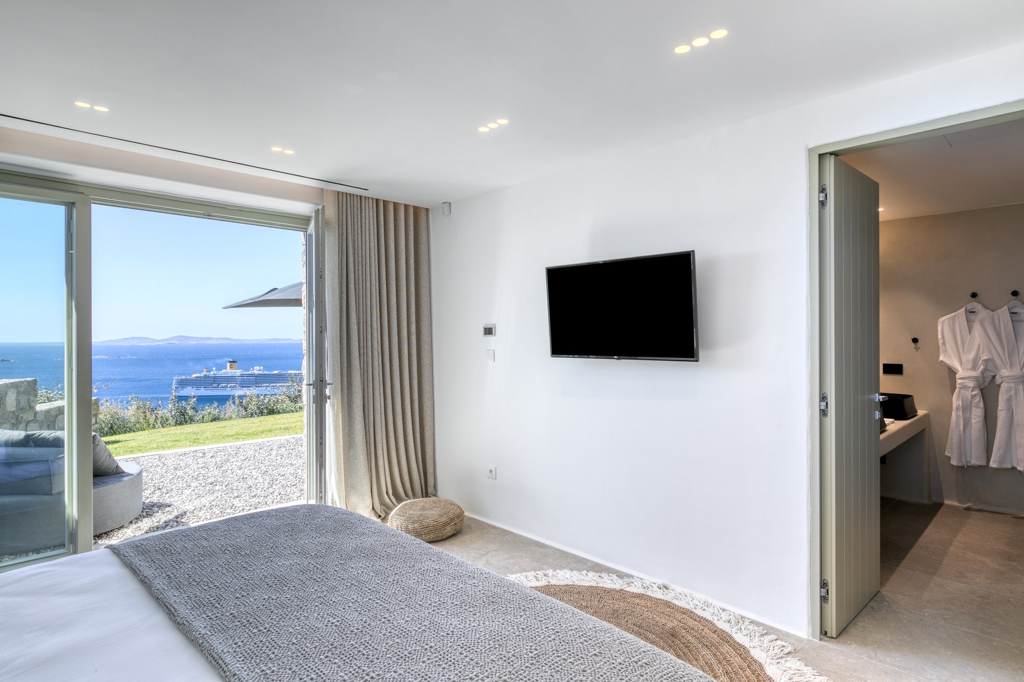 Modern bedroom with ocean view, bed, TV, and open door to bathroom.