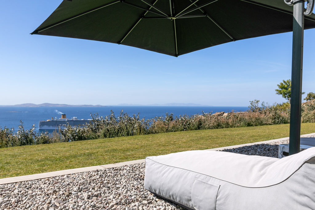 Outdoor lounge chair under a large umbrella with a scenic view of the ocean and a ship.