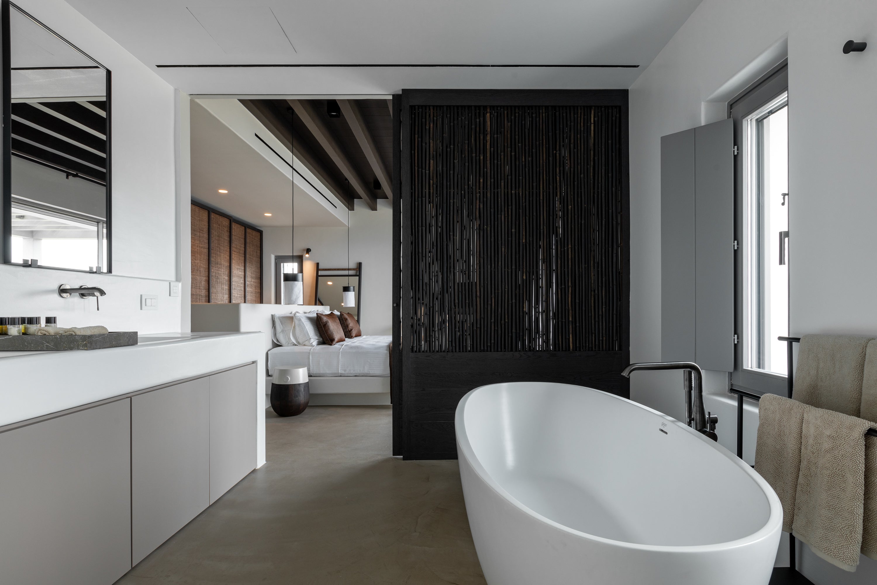 Modern bathroom with a freestanding bathtub, sink, and bed in the background.