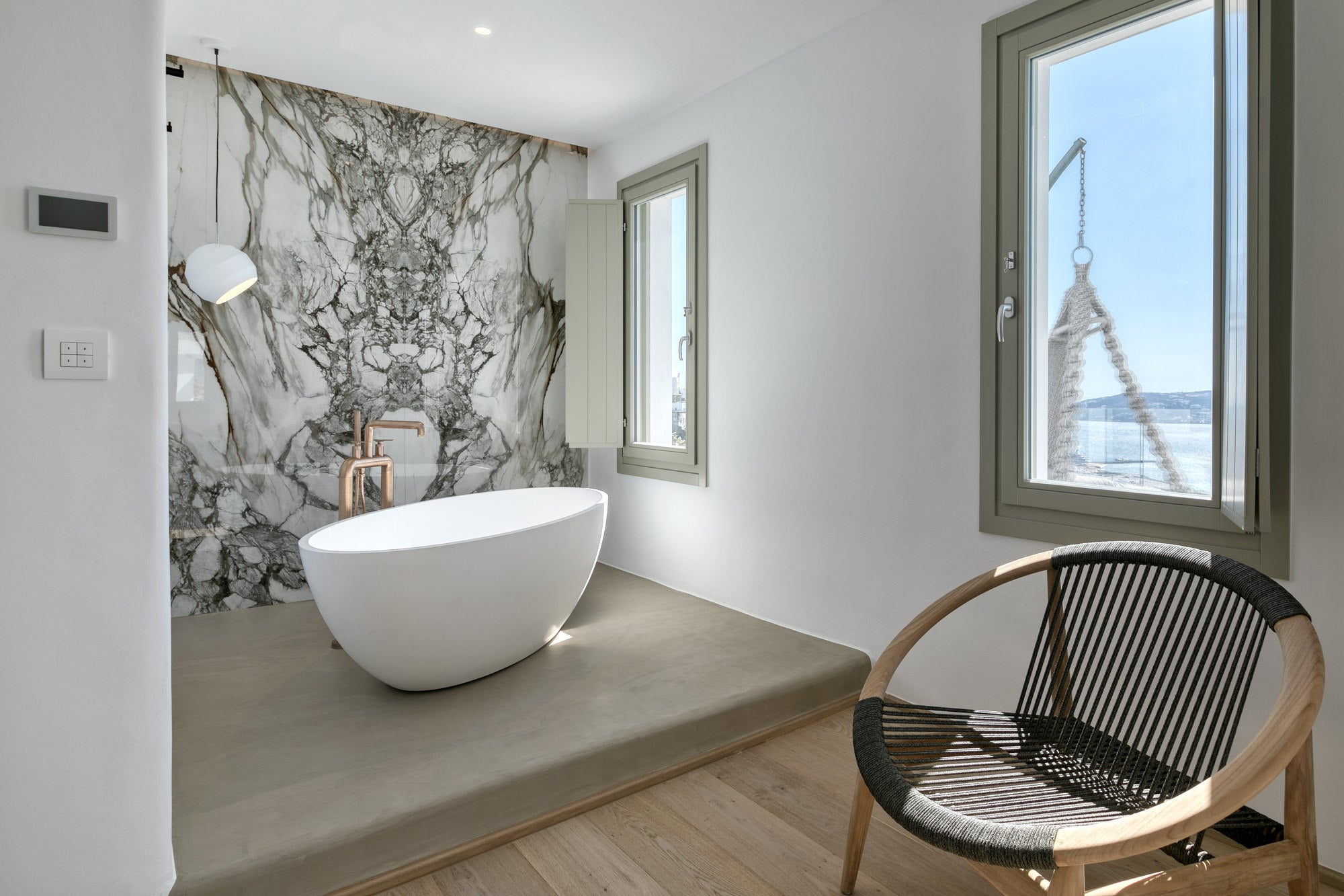 Modern bathroom with a freestanding bathtub, marble wall, and a chair with a view of the ocean.