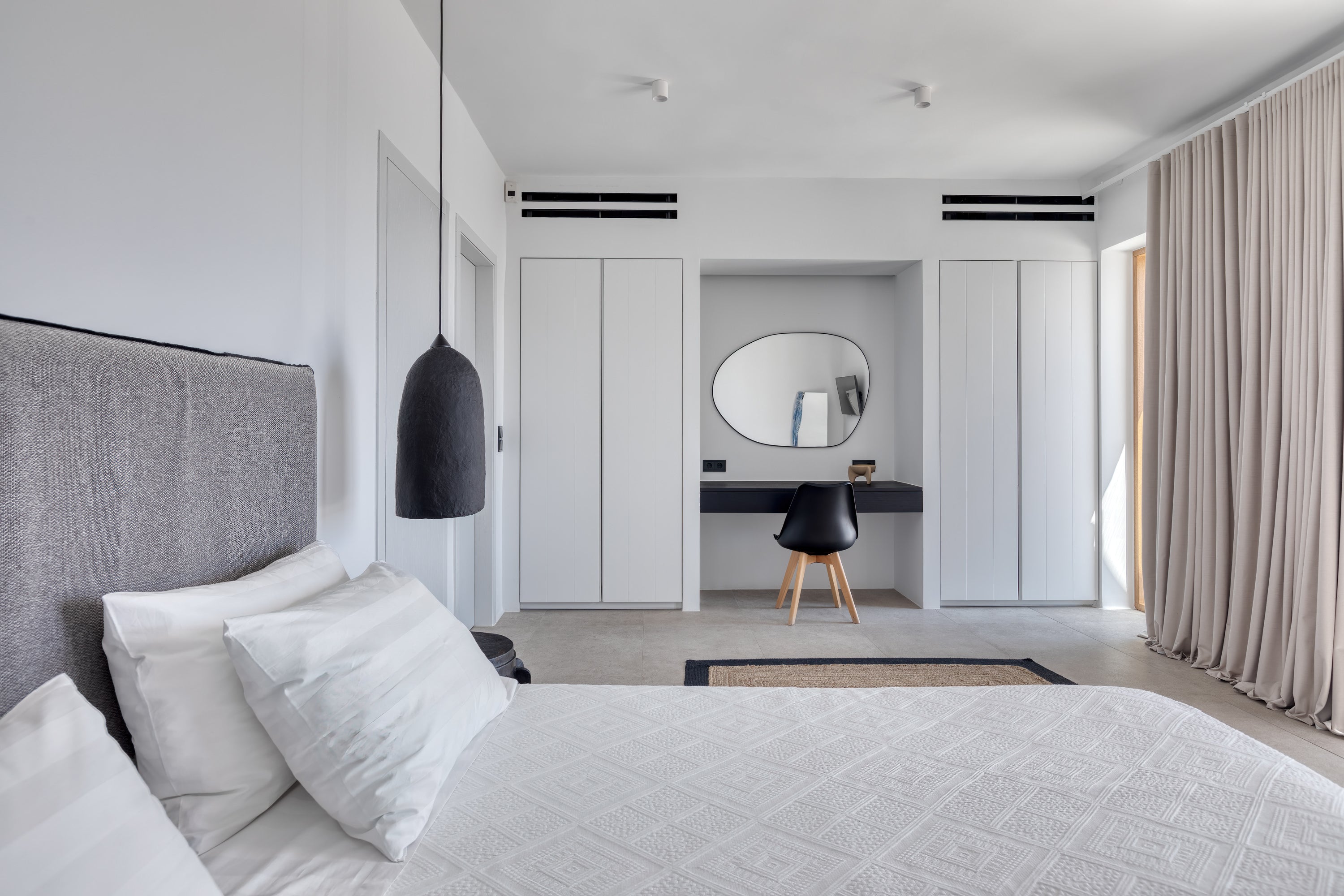 Modern bedroom with gray headboard, white bedding, and a mirror reflecting a desk area.