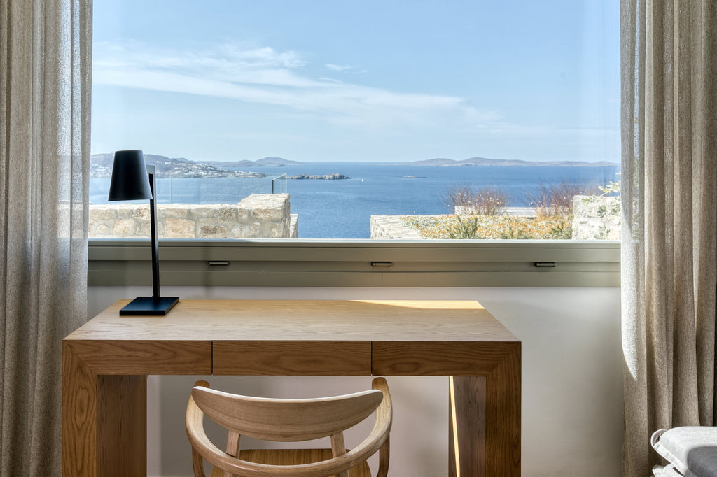 Wooden desk with chair in a room with a large window overlooking water and buildings.