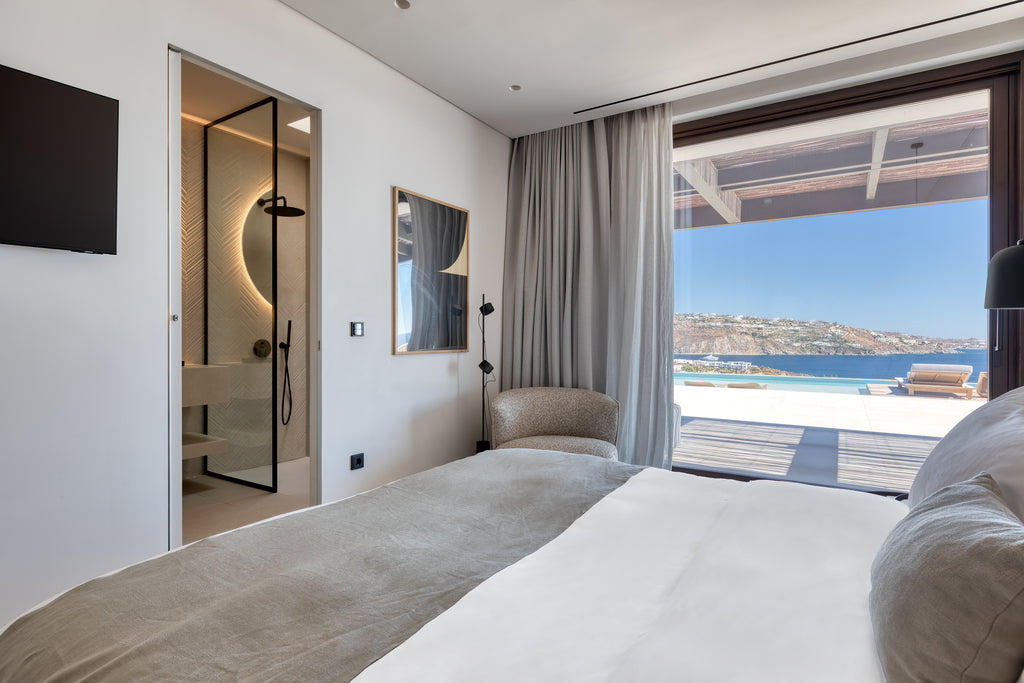 Modern bedroom with a large bed, a view of the sea through a window, and a bathroom visible.