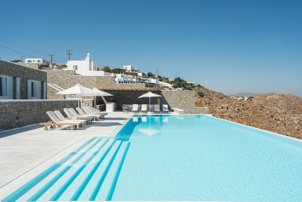 Pool area with lounge chairs and umbrellas in a scenic location with white buildings and blue sky.