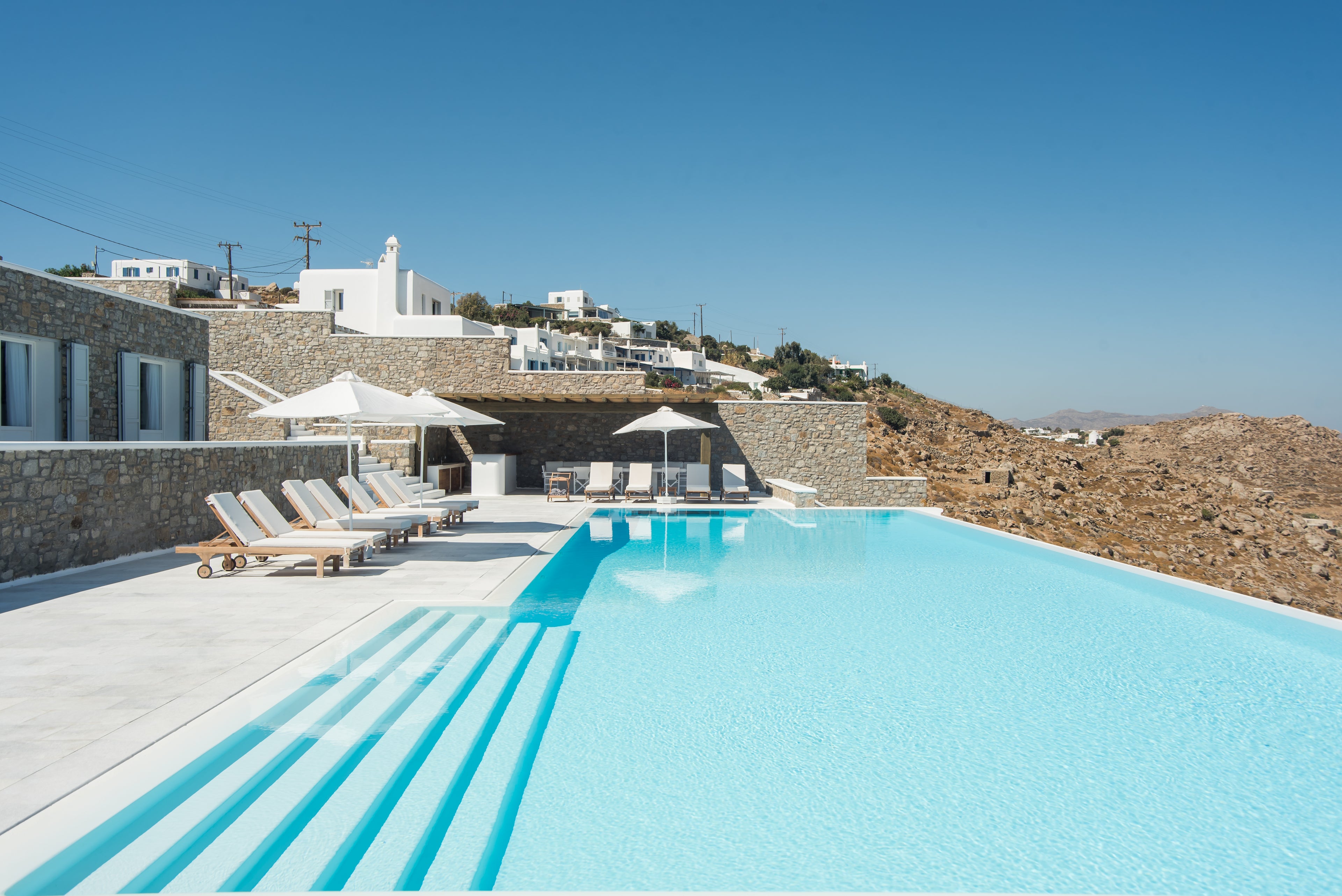 Pool area with lounge chairs and umbrellas in a scenic location with white buildings and blue sky.
