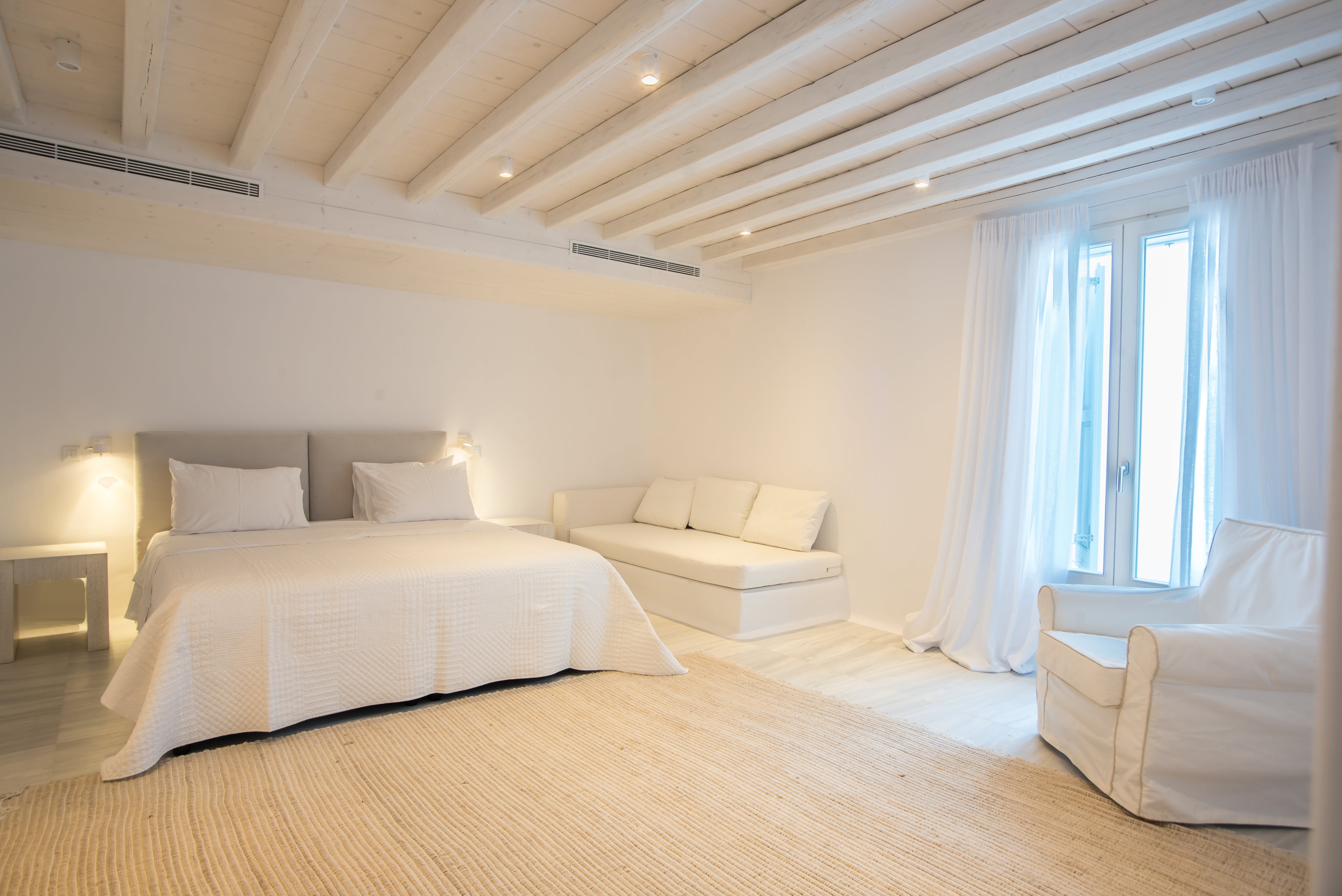 Modern bedroom with white bedding, light wood flooring, and large windows.