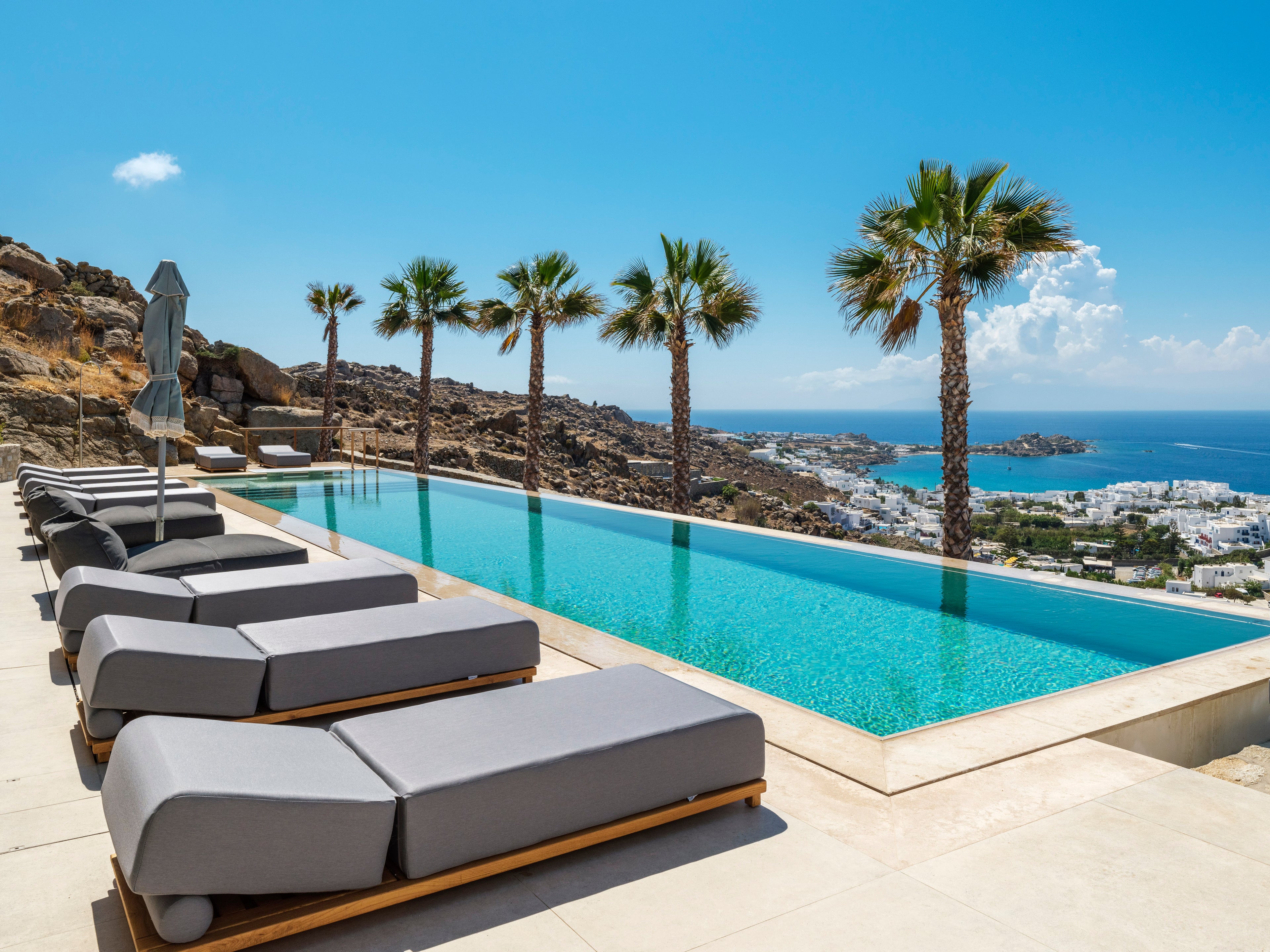Row of lounge chairs by a pool with a scenic view of the ocean and palm trees.