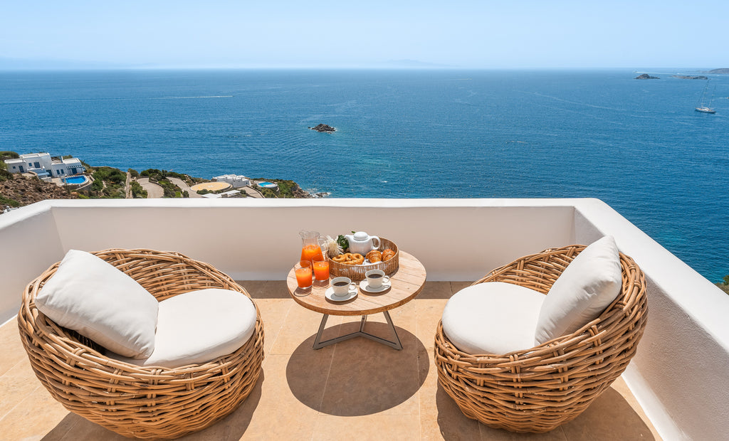 Two wicker chairs with white cushions on a balcony overlooking the ocean.