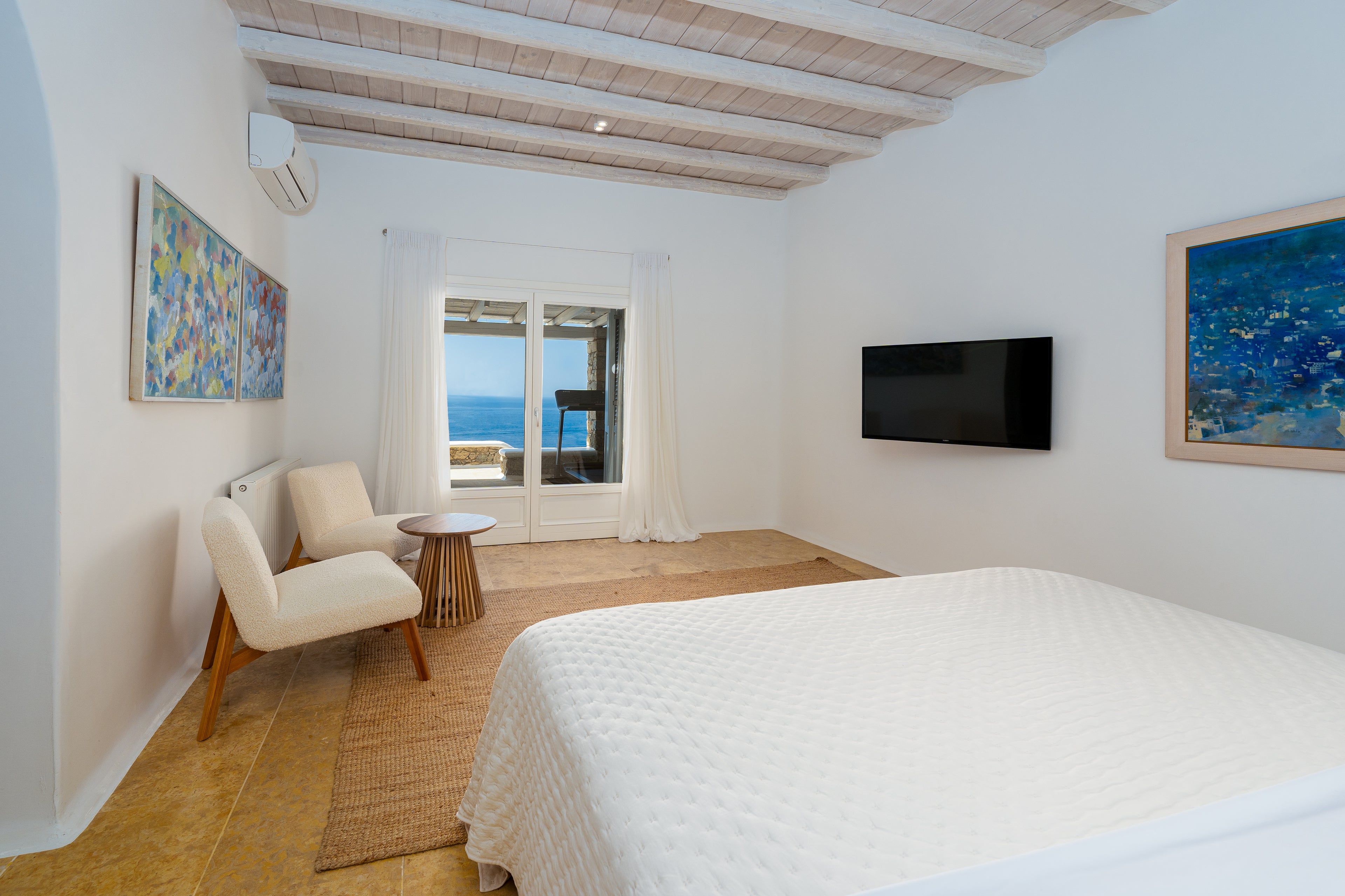 Modern bedroom with a bed, chair, television, and large windows overlooking the ocean.