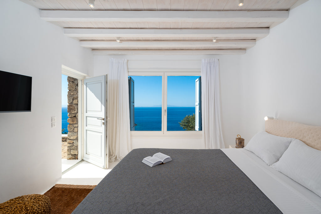 Modern bedroom with ocean view through large windows, featuring a bed and television.