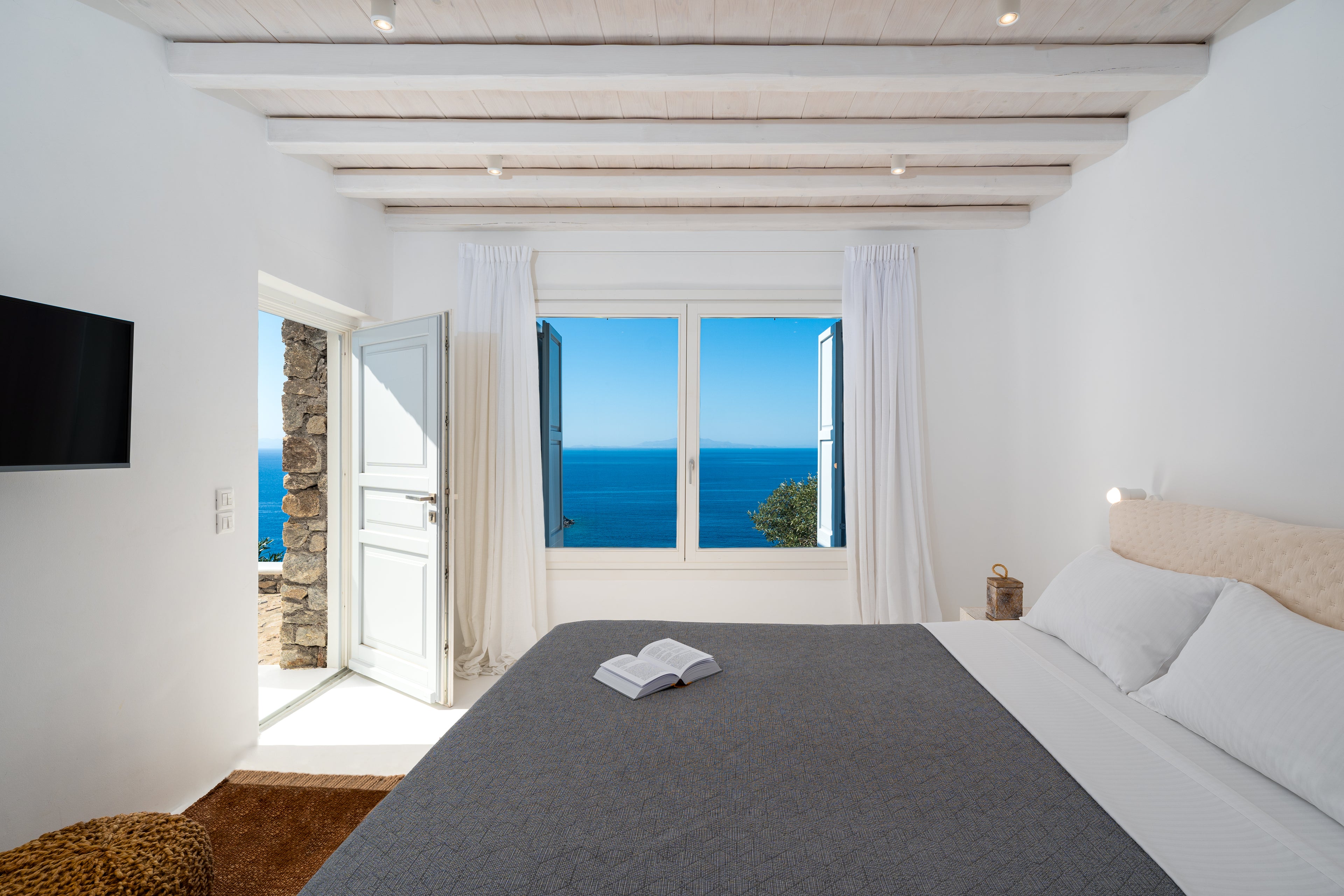 Modern bedroom with ocean view through large windows, featuring a bed and television.