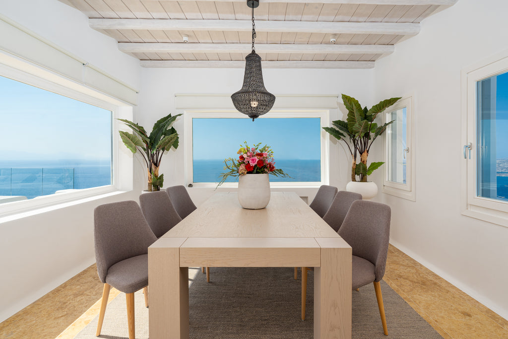 Dining area with a table and chairs in a room with large windows overlooking the ocean.