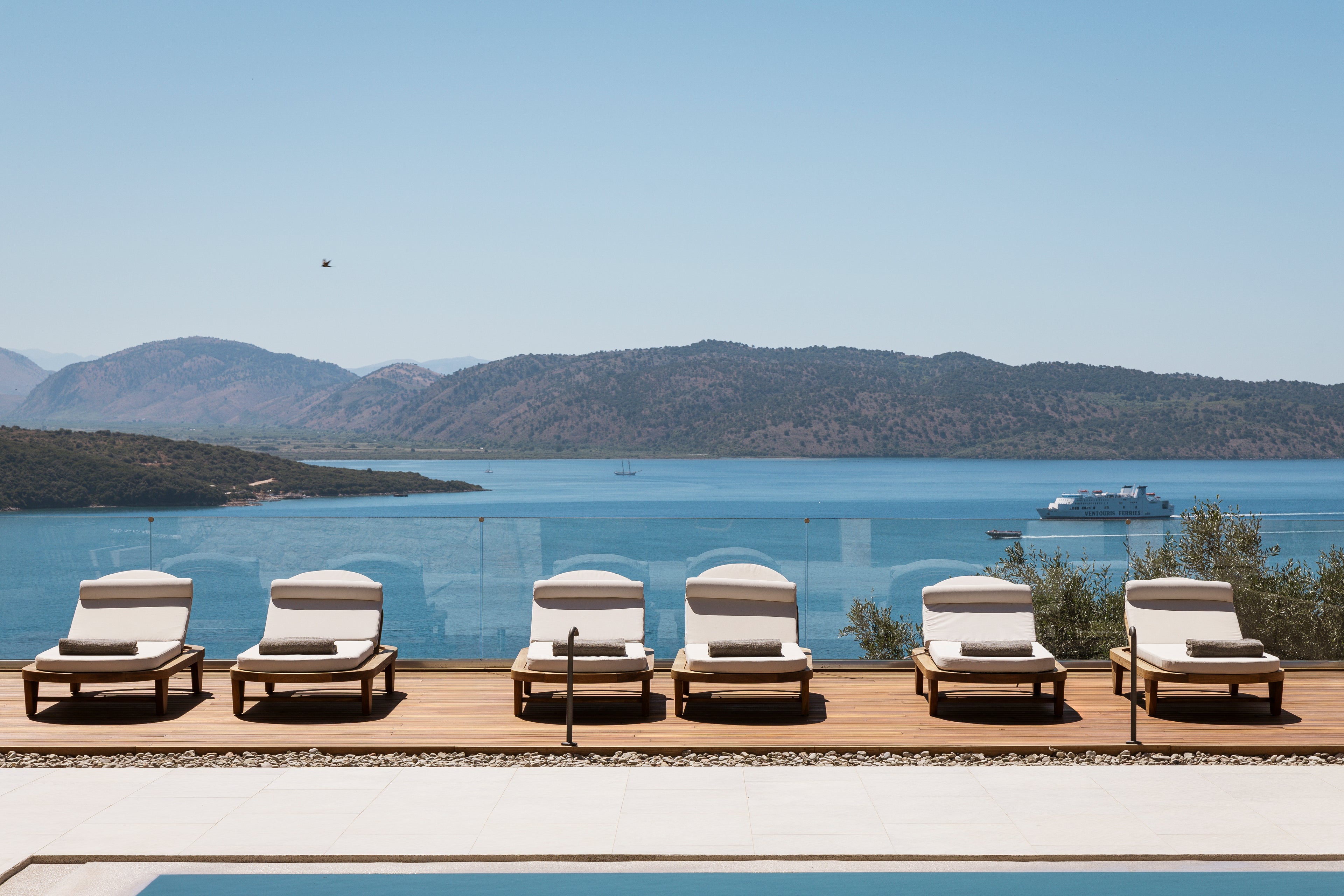 Row of lounge chairs by a pool with a scenic view of water and mountains.