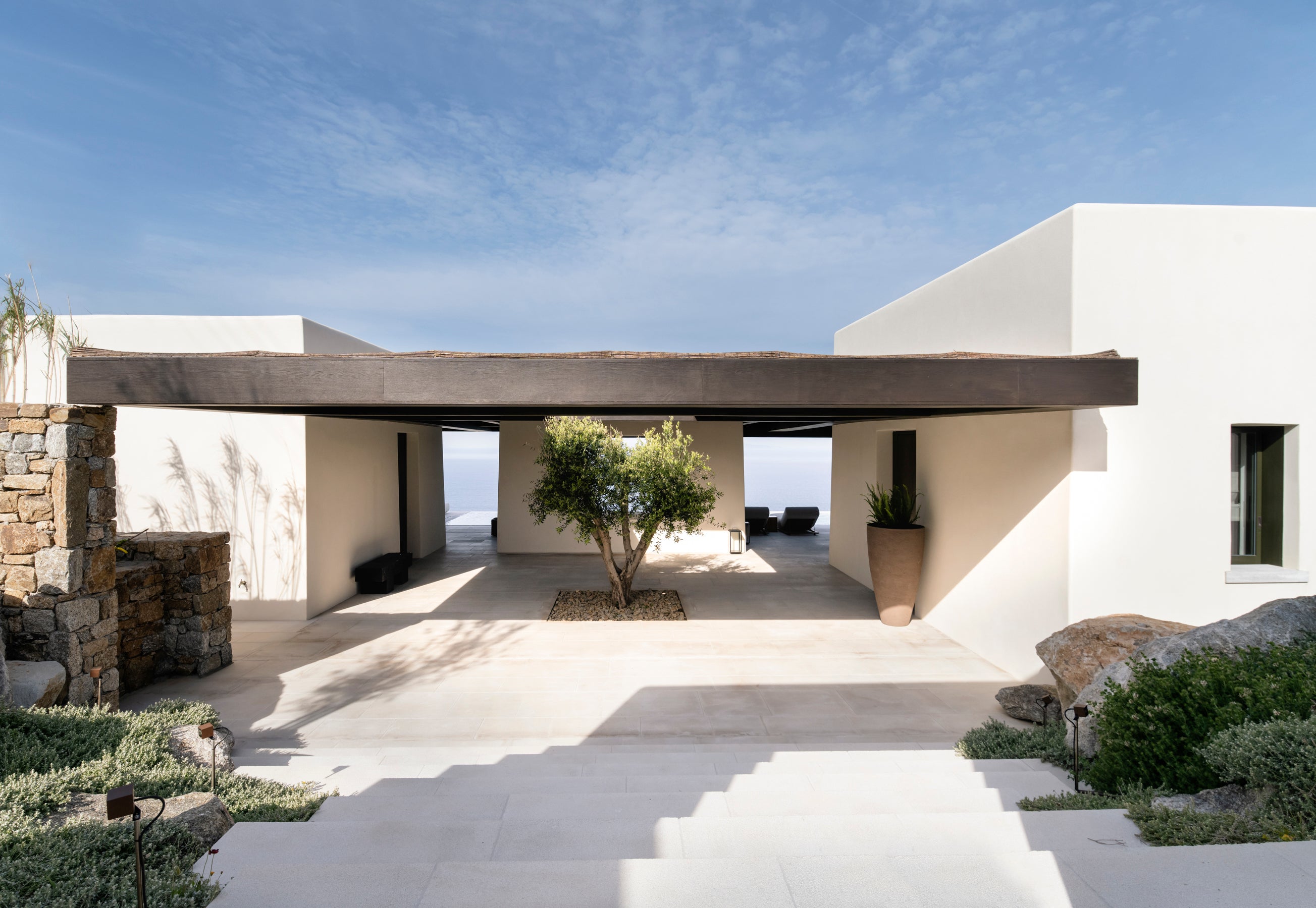 Modern white house with a stone wall and outdoor seating area under a clear blue sky.