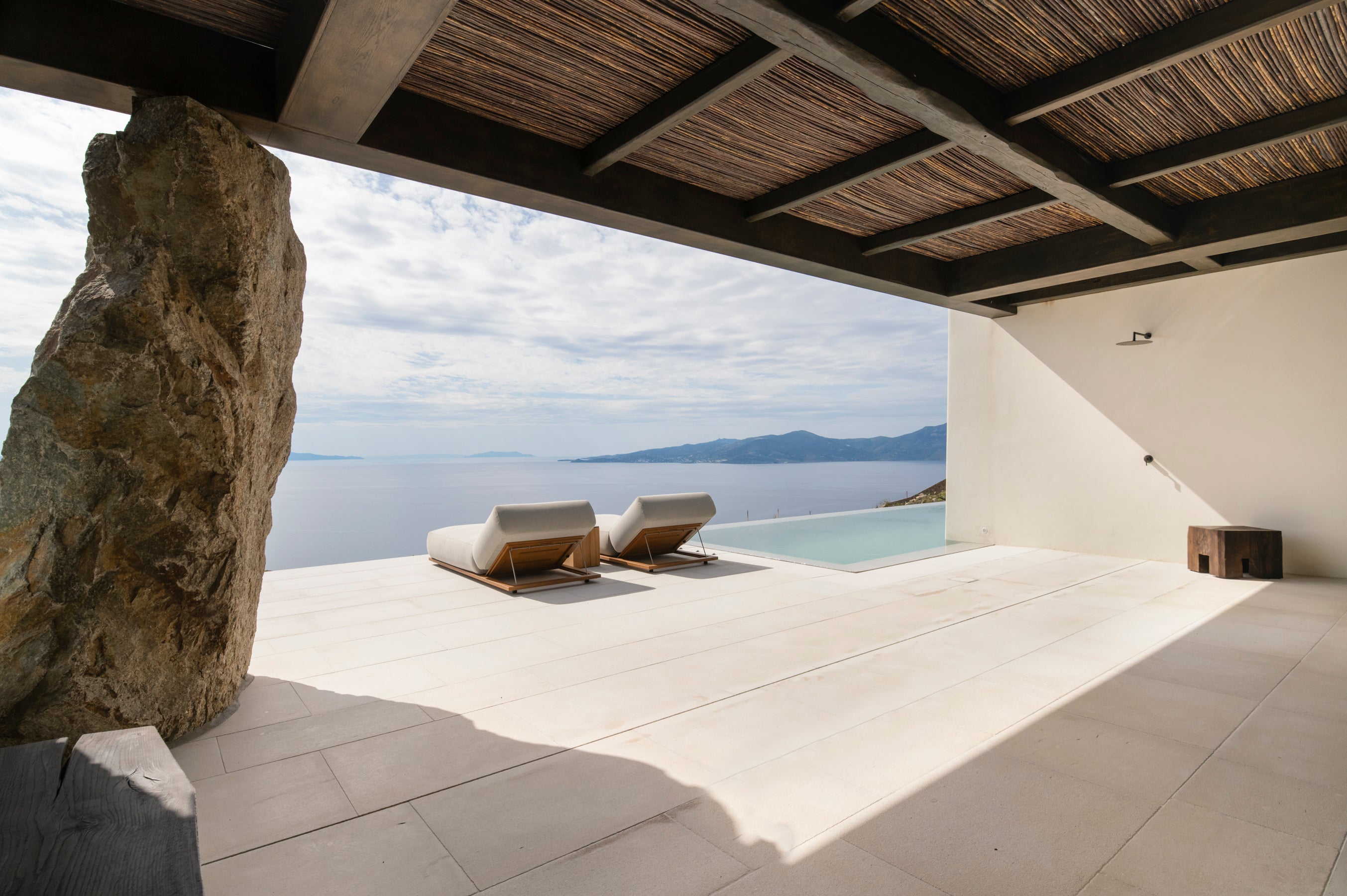 Modern outdoor patio with lounge chairs, stone wall, and pool overlooking a scenic landscape.