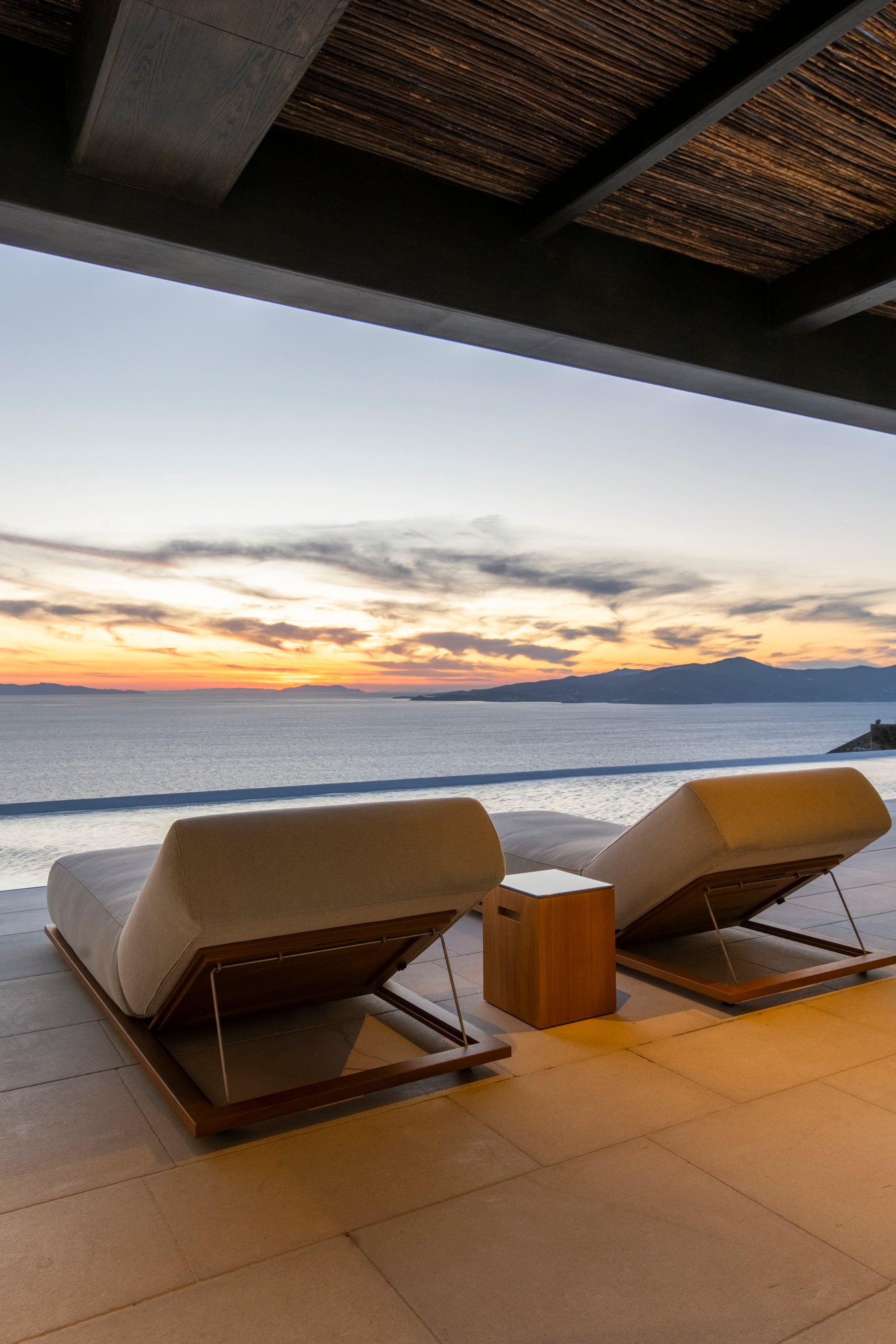 Two lounge chairs on a patio overlooking a sunset over water.