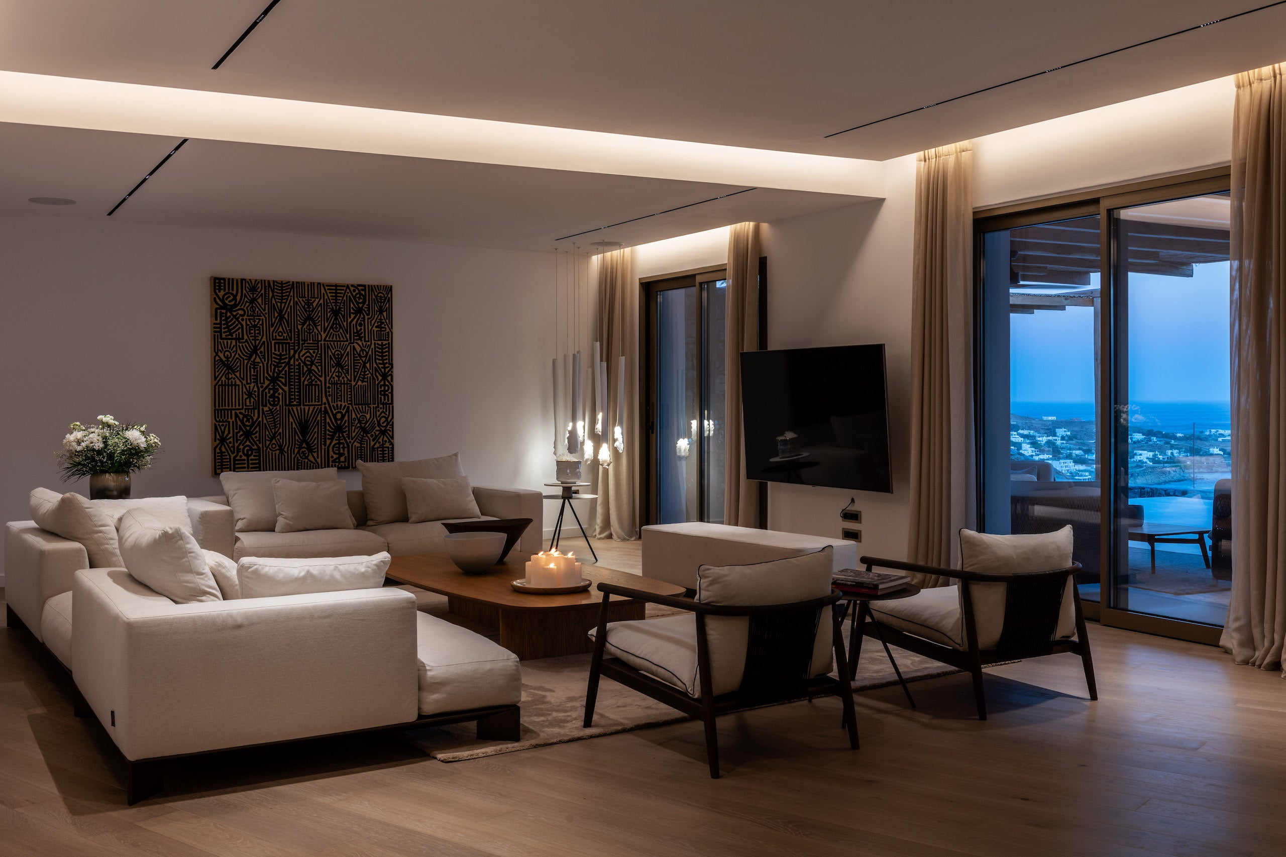 Modern living room with white furniture, a TV, and large windows overlooking a cityscape.