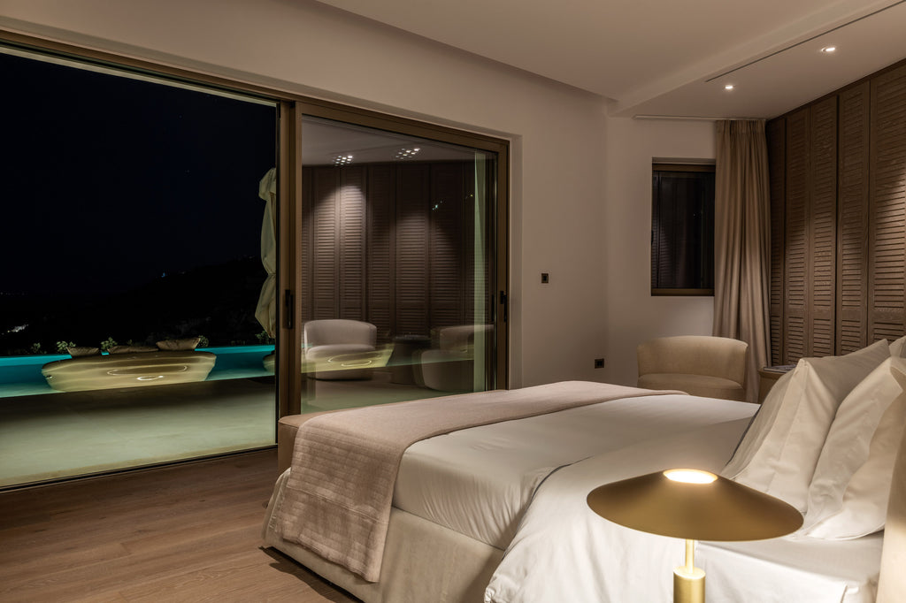 Modern bedroom with a large bed, lamp, and view of a pool at night.