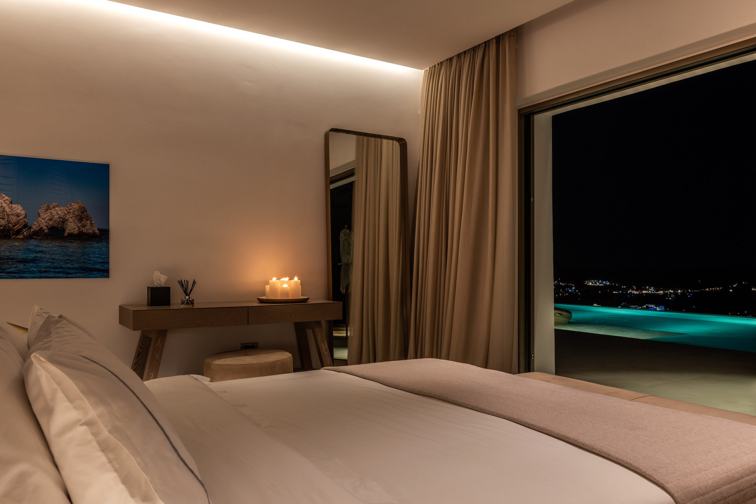 Modern bedroom with bed, desk, and large window overlooking a pool at night.